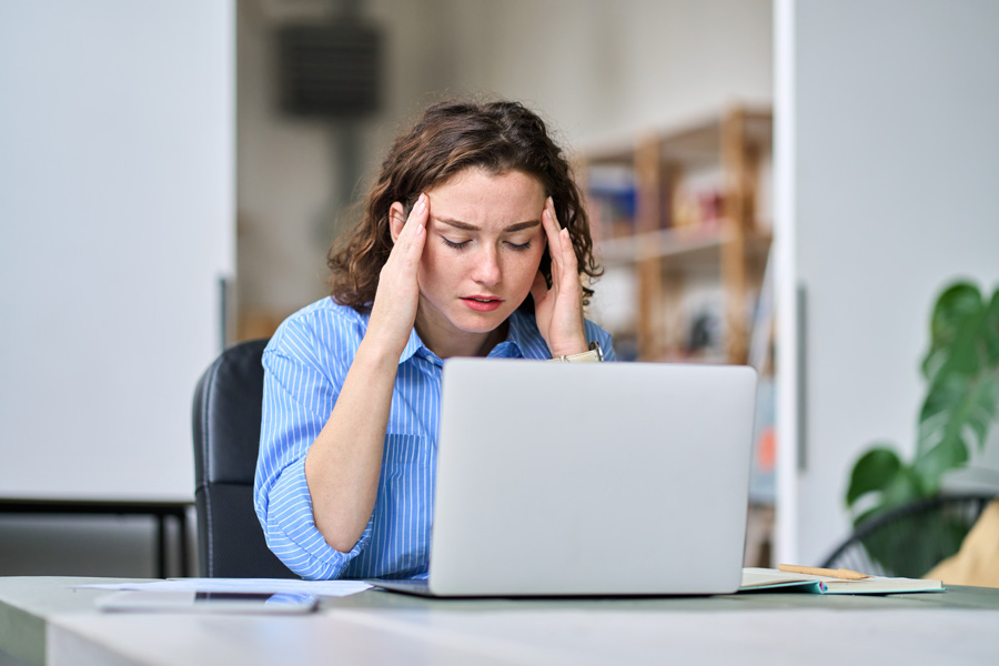 Woman in a blue striped shirt sitting at a desk with a laptop, holding her head with both hands and looking stressed or tired.